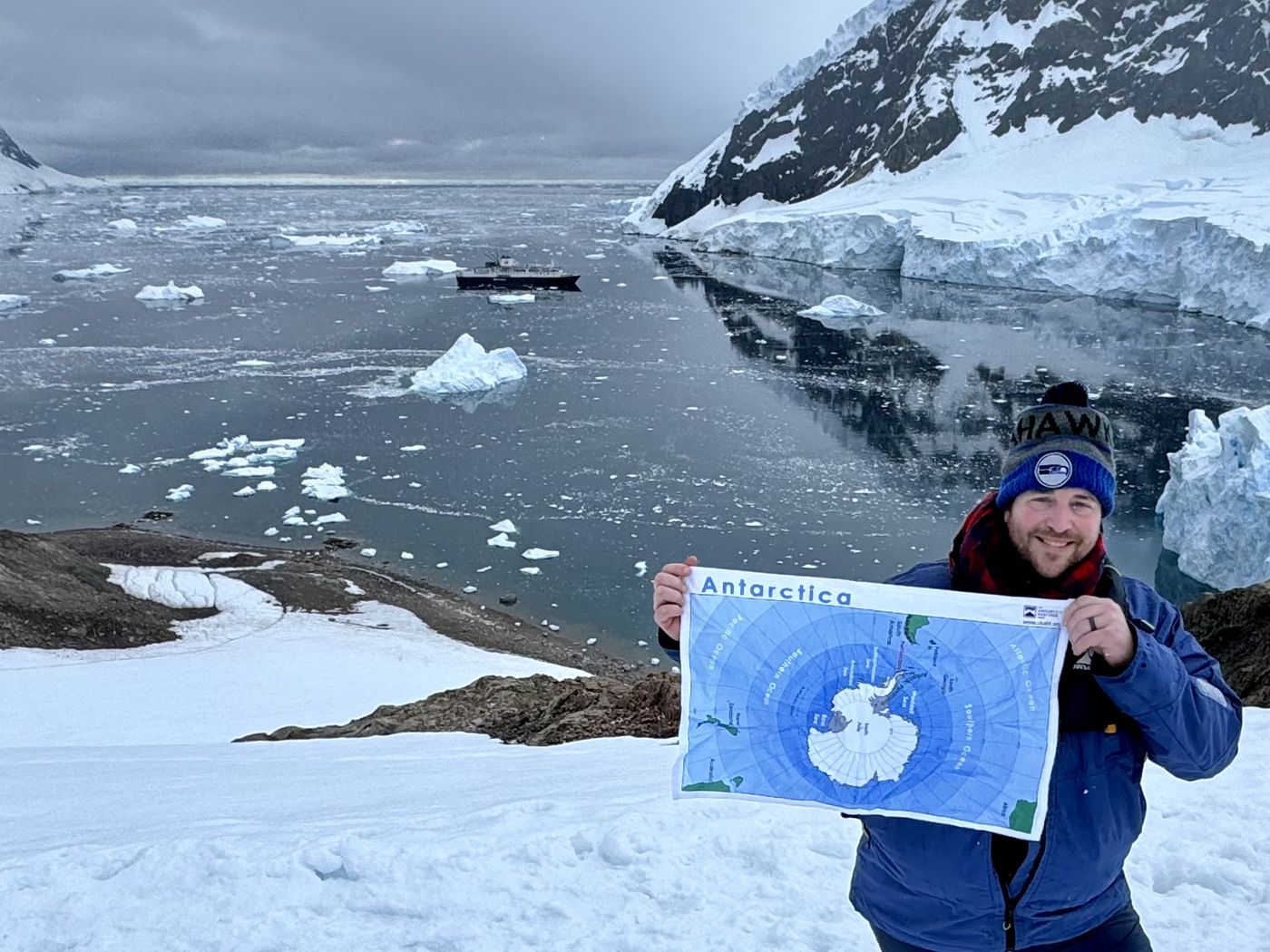 Jacob Schroeder standing in the snow in Antarctica, holding up a map of the continent, with a ship and icebergs in the bay behind him.